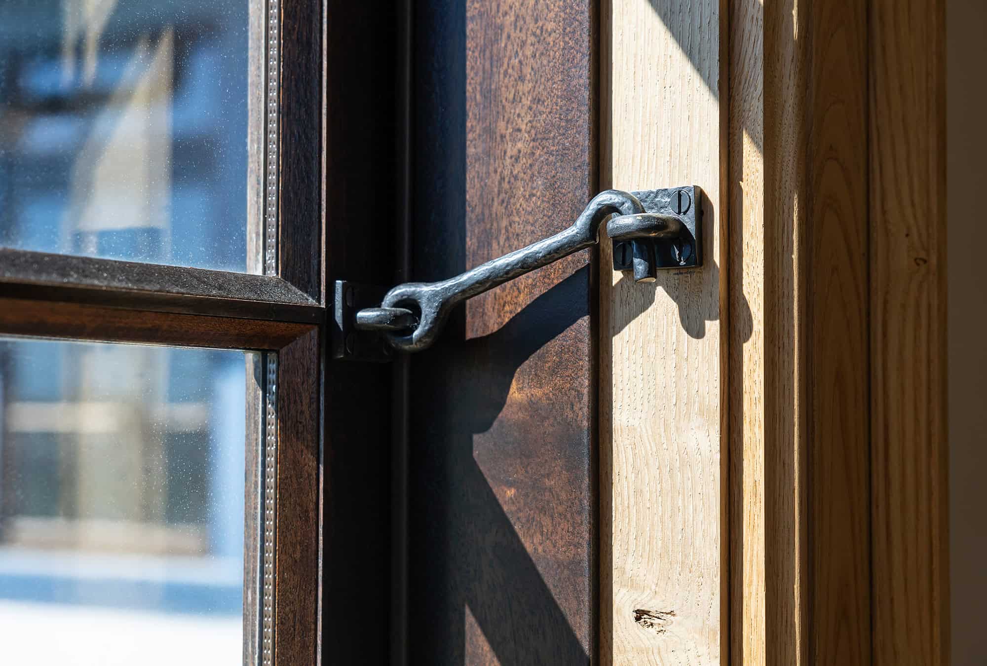 A close-up of a window secured with a metal latch hook fastened to a wooden frame in a luxury horse barn. Sunlight streams through the glass, casting elegant shadows on the wood.