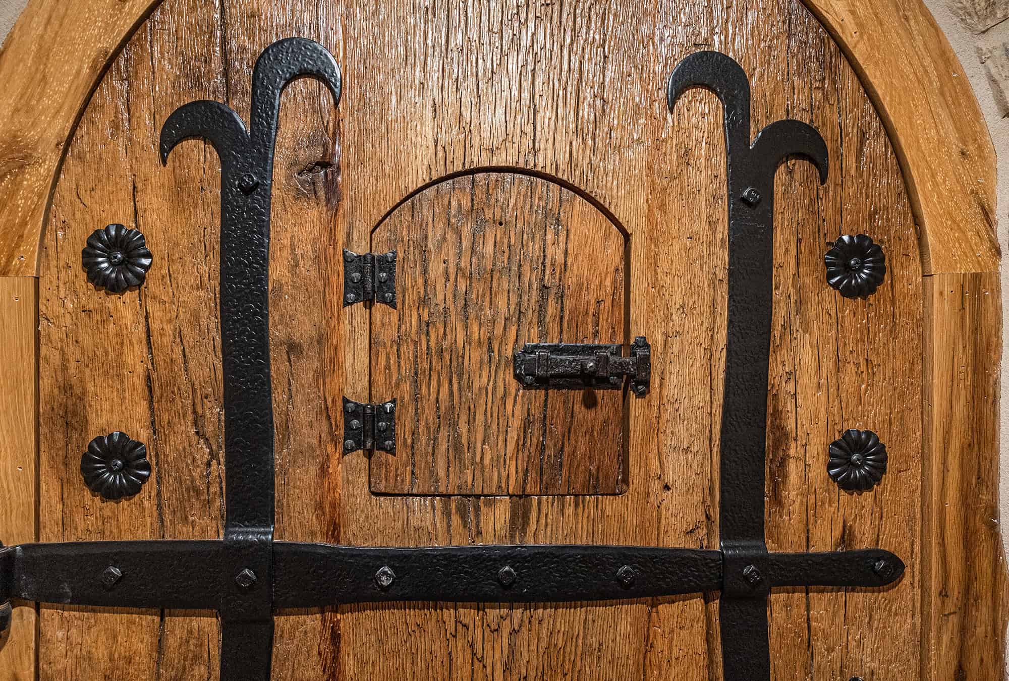 A close-up of a rustic wooden door with ornate black metal hinges and a small rectangular hatch secured by a sliding bolt in the center—perfect for adding character to any luxury horse barn. The door has a rough texture and decorative metal accents.