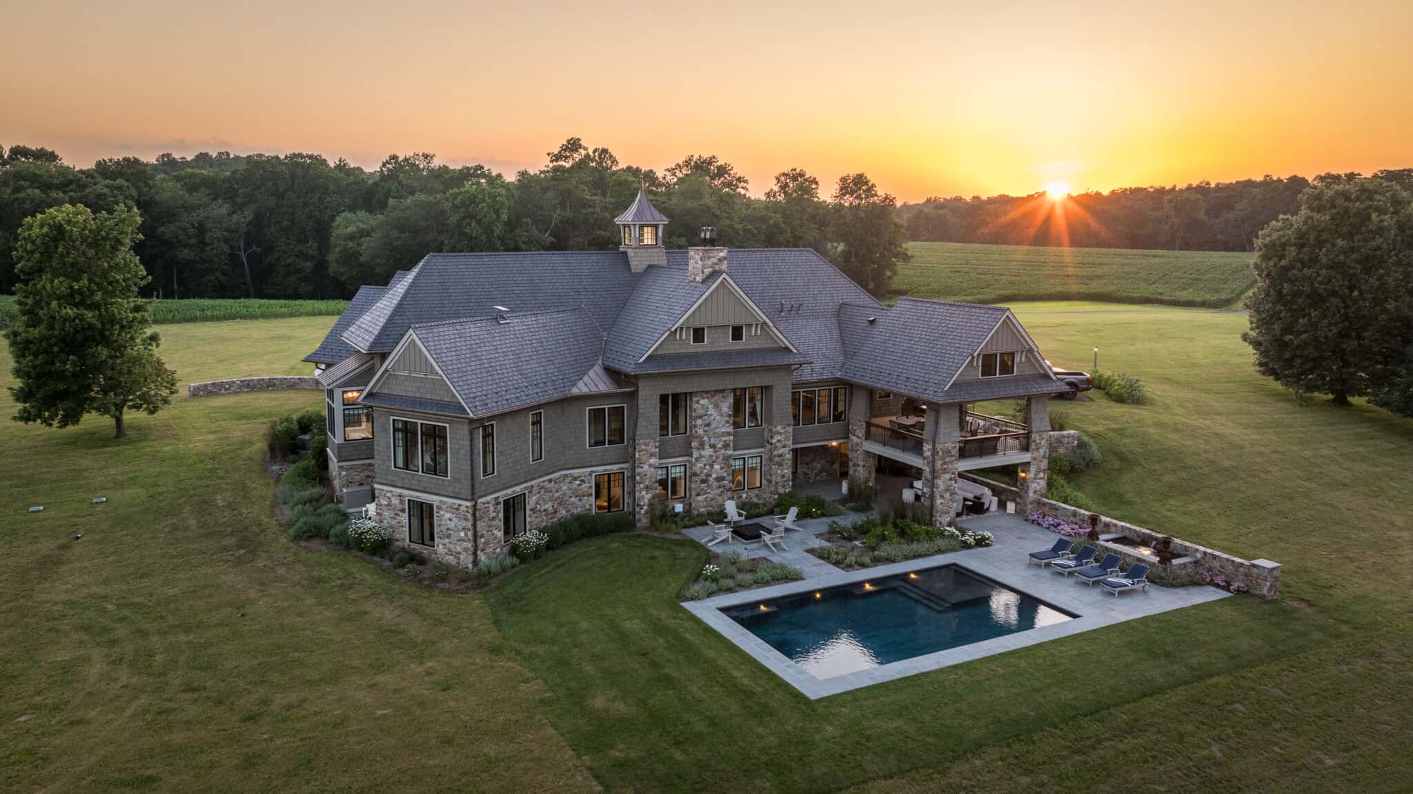 Large, luxurious house with stone accents, multiple gables, and a pool in the backyard, set on a spacious green lawn with trees and fields under a sunset sky—complete with a nearby luxury horse barn for an elegant country lifestyle.