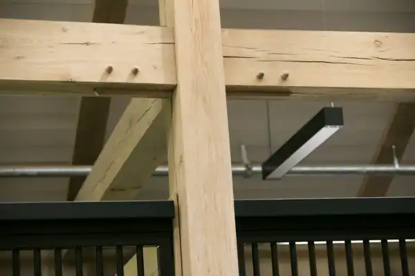 Close-up of intersecting wooden beams in a luxury horse barn’s interior, with visible wooden pegs, part of a black railing below, a modern rectangular light fixture, and exposed ductwork in the background.