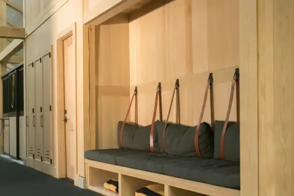 A built-in wooden bench with gray cushions and brown leather straps is set into a wall in this luxury home. Open cubbies below hold folded blankets, with light wood paneling and black flooring adding to the sophisticated vibe.