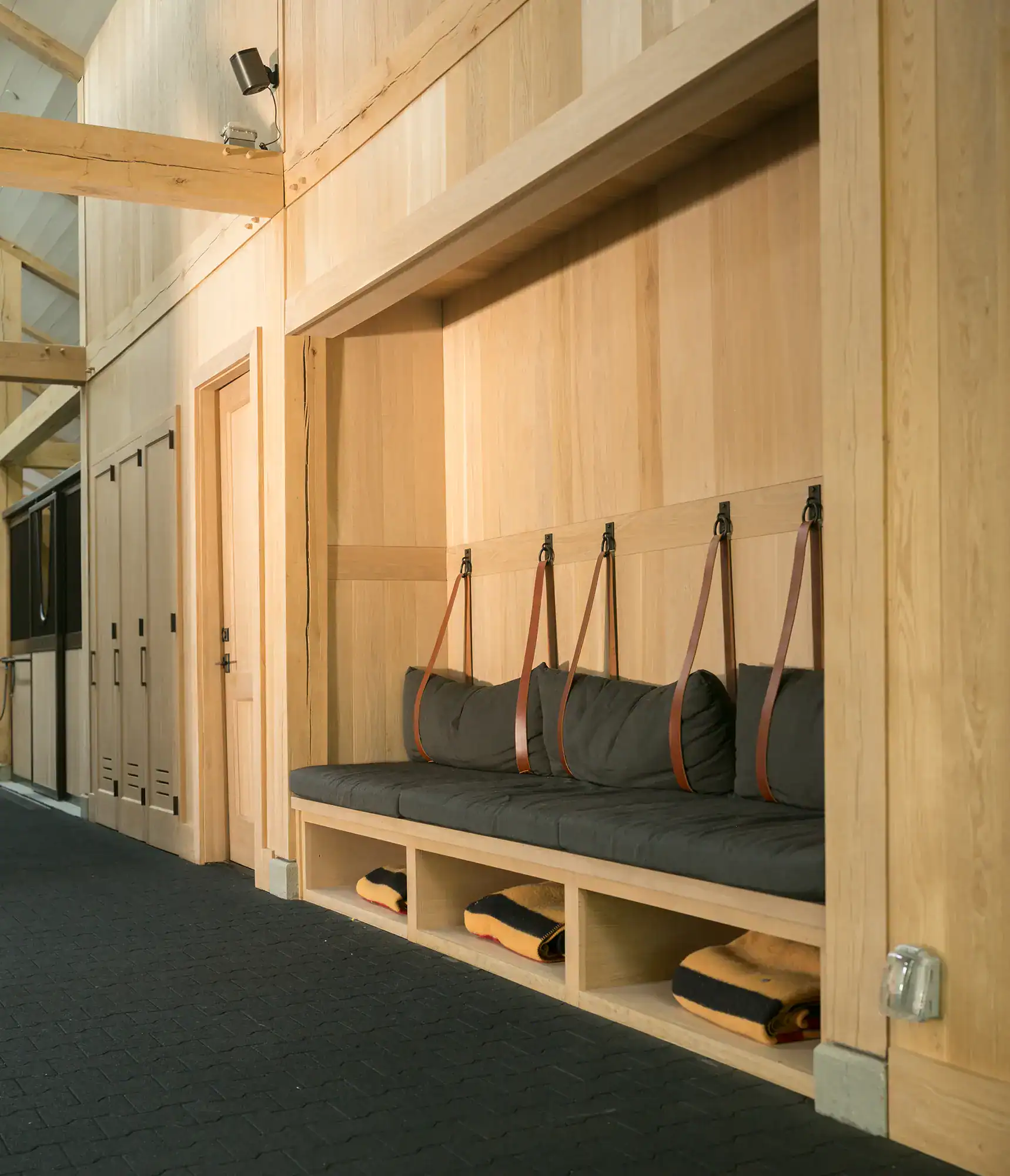 A built-in wooden bench with gray cushions and brown leather straps is set into a wall in this luxury home. Open cubbies below hold folded blankets, with light wood paneling and black flooring adding to the sophisticated vibe.