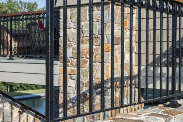 A decorative black metal railing with circular accents sits atop a stone wall, framing the view of a luxury home and trees in the background.