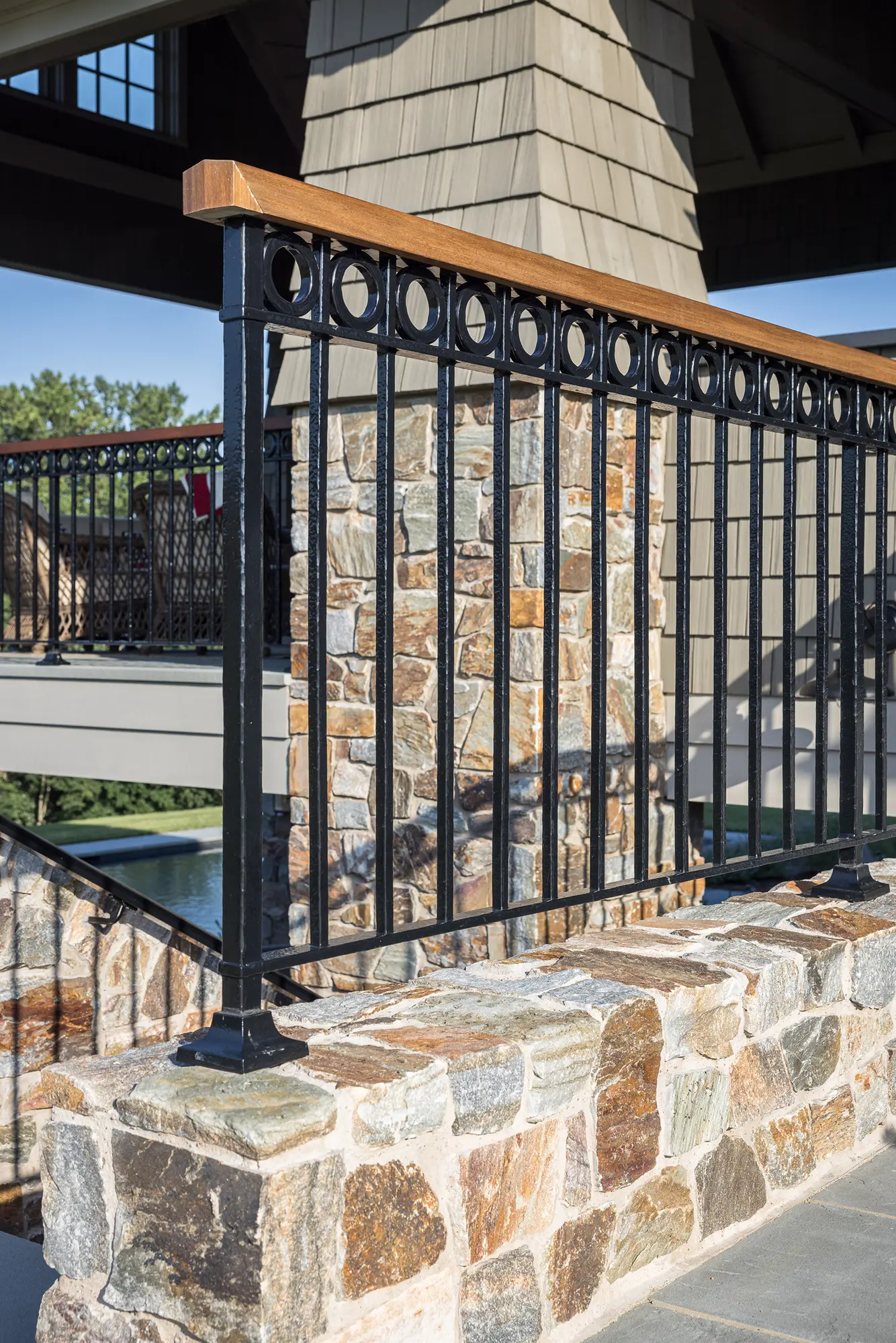 A decorative black metal railing with circular accents sits atop a stone wall, framing the view of a luxury home and trees in the background.