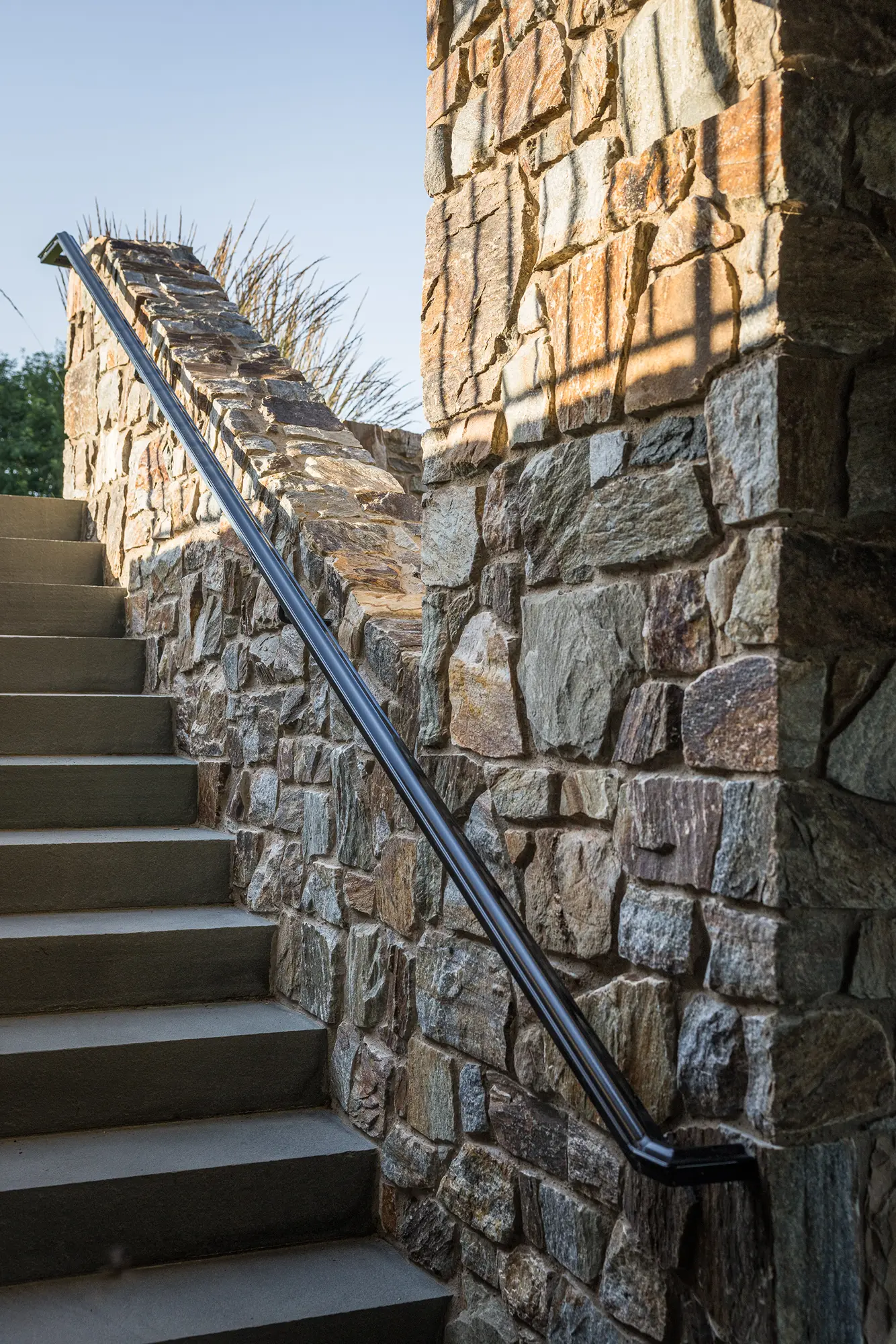 Stone stairway with a smooth metal handrail lines the wall of a luxury home, its irregularly shaped stones adding elegance as sunlight casts shadows on the surface and stairs lead upward.