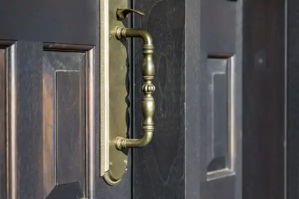 A close-up of an ornate brass door handle on a dark wooden door with carved panels inside a luxury horse barn. Sunlight highlights the details of the handle and wood grain, adding elegance to the barn’s refined setting.