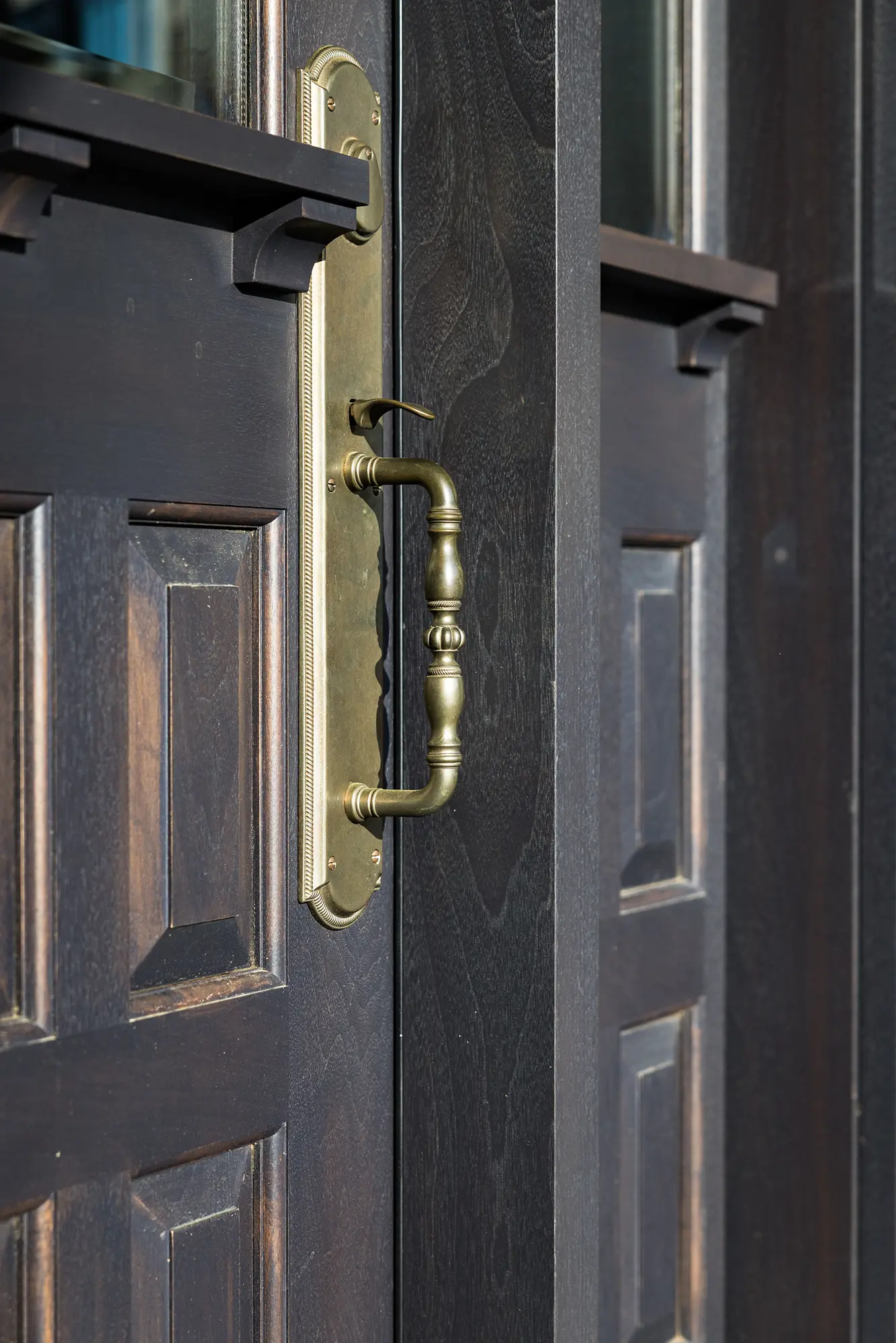 A close-up of an ornate brass door handle on a dark wooden door with carved panels inside a luxury horse barn. Sunlight highlights the details of the handle and wood grain, adding elegance to the barn’s refined setting.