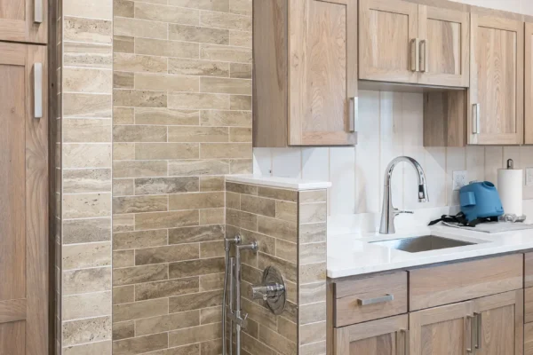 A modern kitchen in a luxury home with light wood cabinets, white countertops, and a small tiled pet washing station built into the cabinetry, featuring a handheld shower head and brown tile.