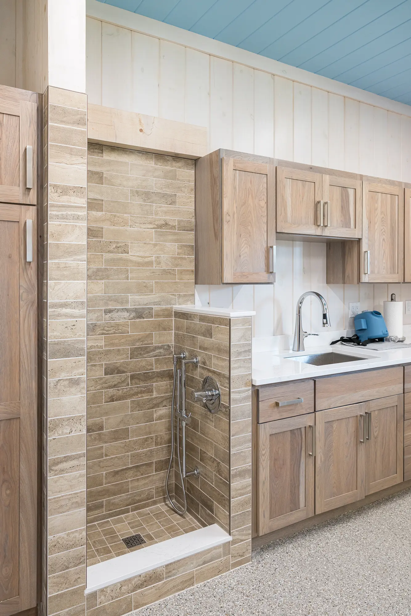 A modern kitchen in a luxury home with light wood cabinets, white countertops, and a small tiled pet washing station built into the cabinetry, featuring a handheld shower head and brown tile.