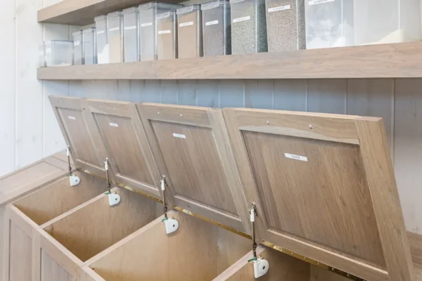 Open wooden bins, one filled with dried leaves, sit below shelves holding clear containers of grains and seeds in a bright, organized bulk food storage area reminiscent of a well-appointed bank barn.