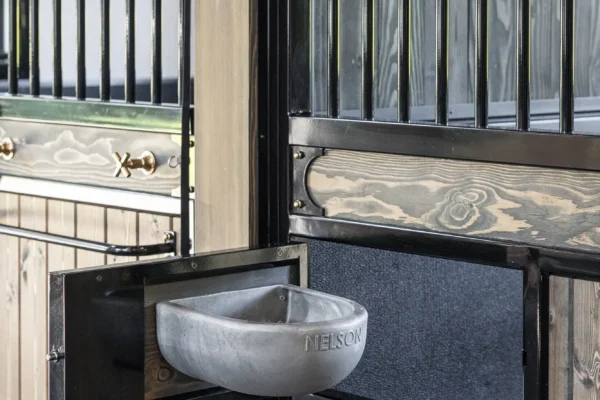 A Nelson metal automatic water bowl is mounted to the side of a wooden and black metal horse stall in a luxury horse barn, with vertical bars and hardware visible in the background.