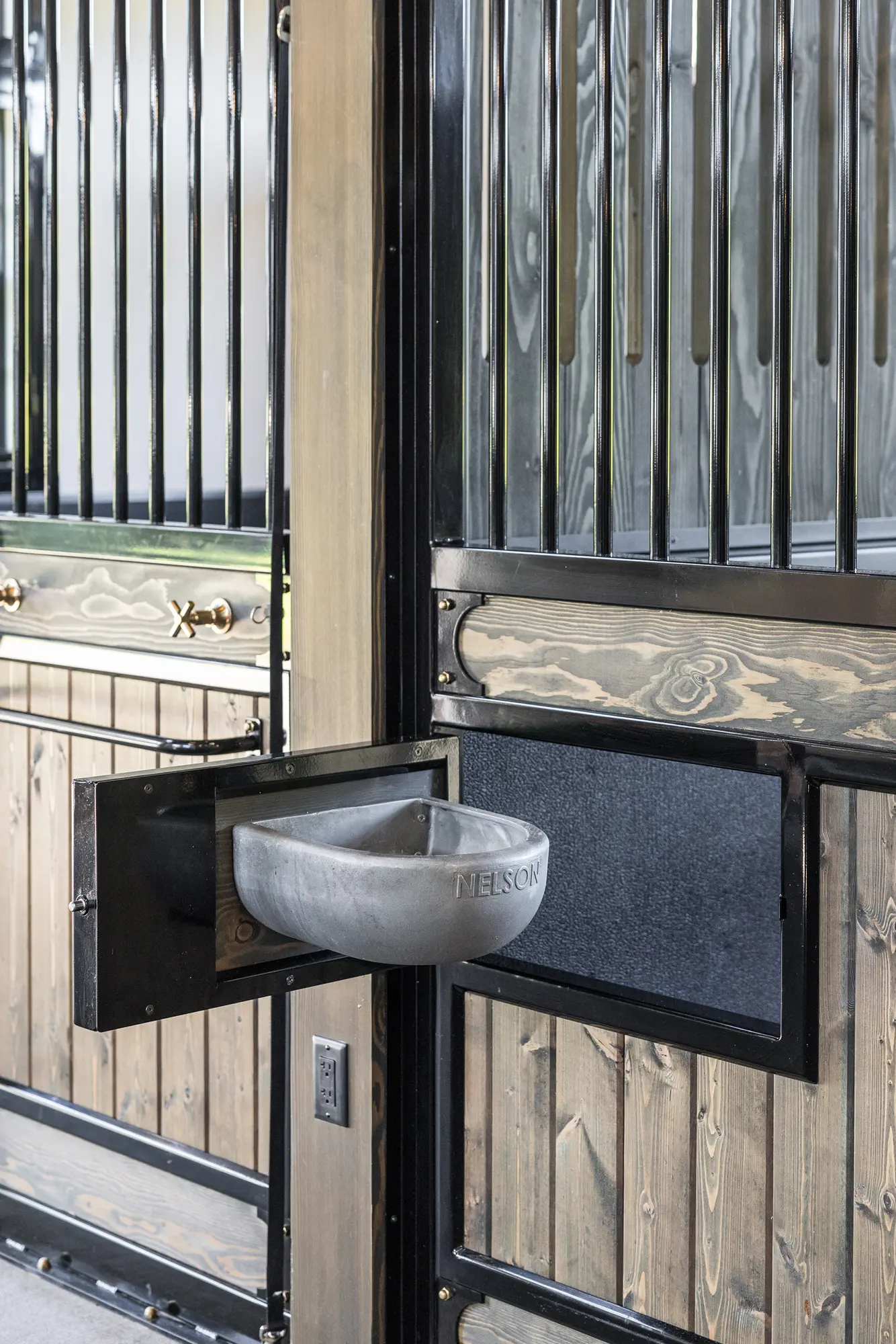 A Nelson metal automatic water bowl is mounted to the side of a wooden and black metal horse stall in a luxury horse barn, with vertical bars and hardware visible in the background.
