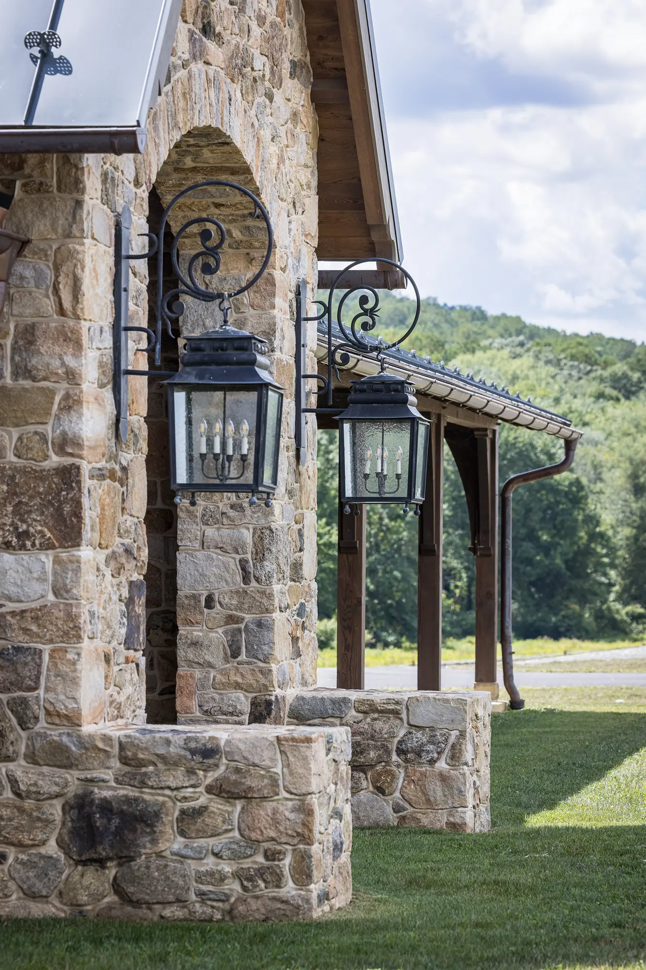 Large black lantern-style sconces mounted on a rustic stone wall outside a luxury home, with green grass, trees, and a cloudy sky in the background.