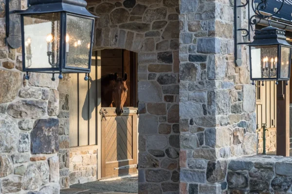 A stone archway leads to a party barn stable with large lanterns on either side. Inside, a brown horse looks out over a half-door. Green grass borders the stone path, adding charm to this luxury home setting.