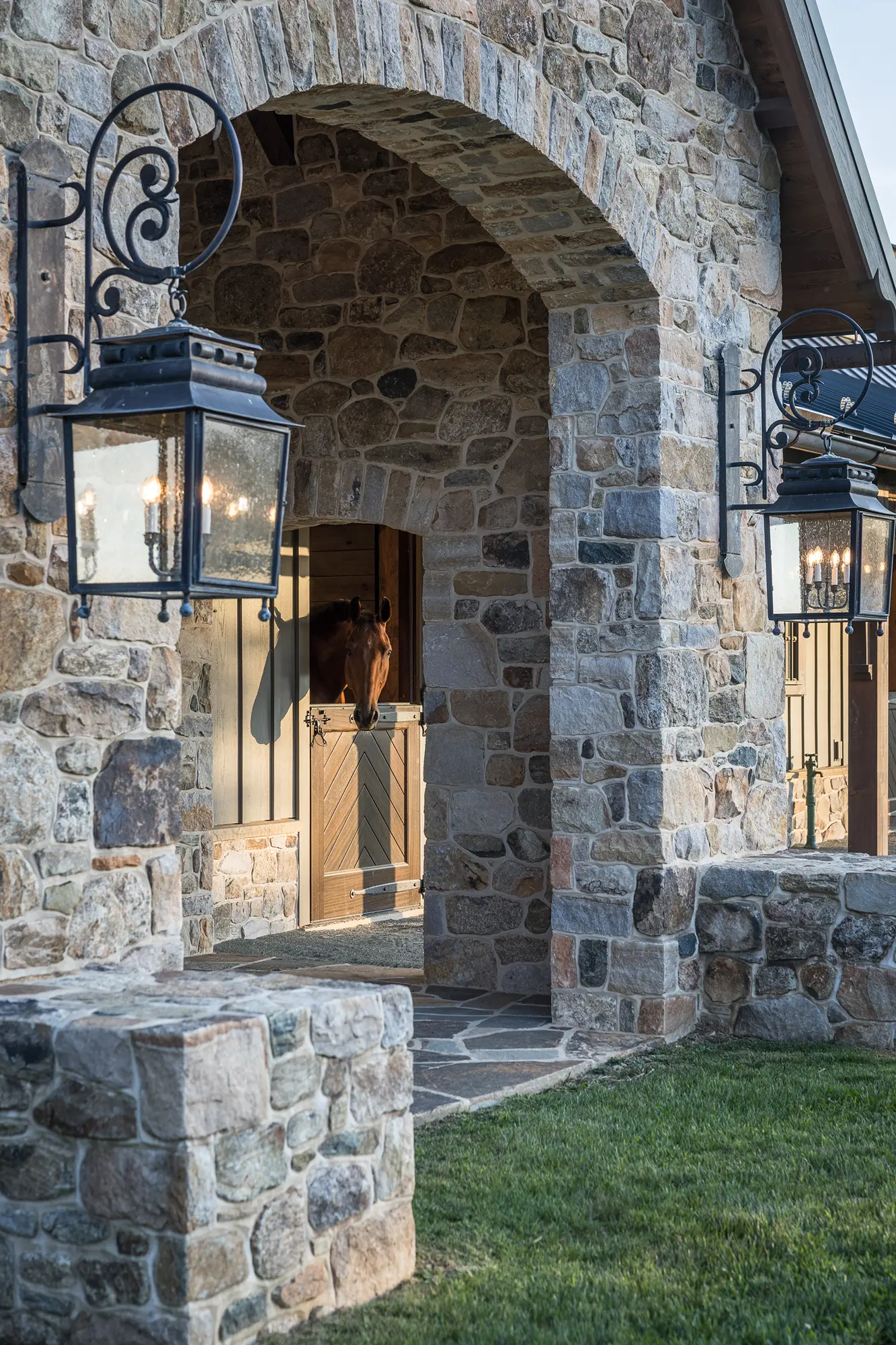 A stone archway leads to a party barn stable with large lanterns on either side. Inside, a brown horse looks out over a half-door. Green grass borders the stone path, adding charm to this luxury home setting.