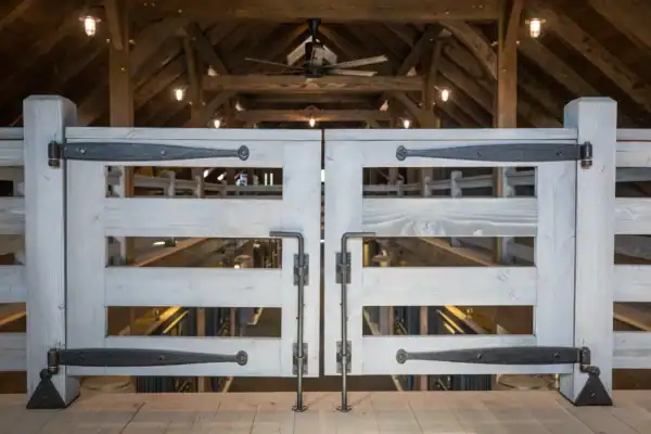 A close-up of double barn doors with large black metal hinges and handles, set inside a party barn with exposed beams, ceiling lights, and a ceiling fan in the background.