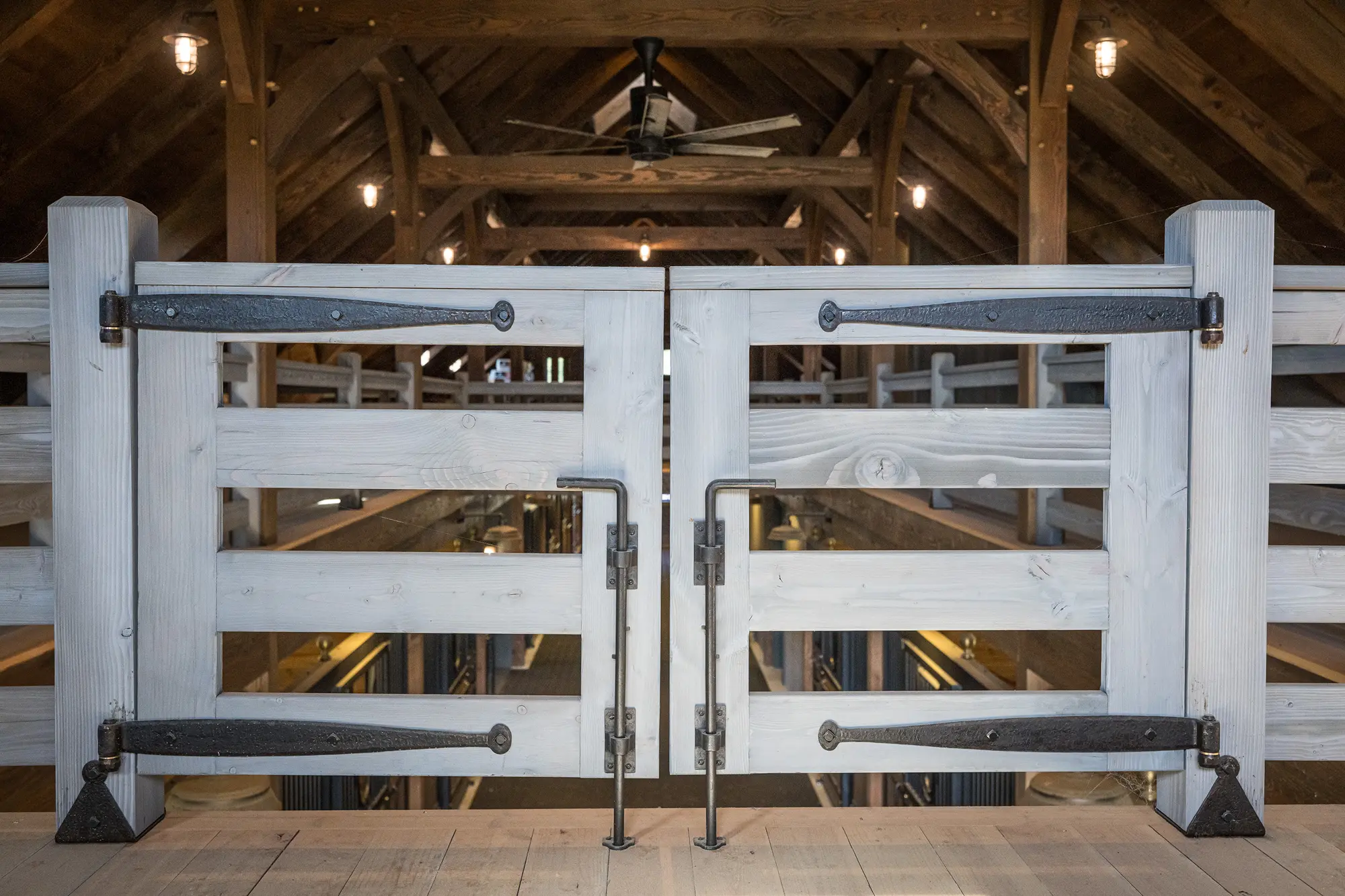 A close-up of double barn doors with large black metal hinges and handles, set inside a party barn with exposed beams, ceiling lights, and a ceiling fan in the background.