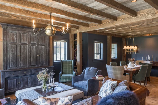 A cozy living and dining area in this luxury home features exposed wooden beams, dark wood cabinets, armchairs, a brown leather sofa, and a dining table under a chandelier. Neutral tones and soft lighting create a warm, inviting party barn ambiance.
