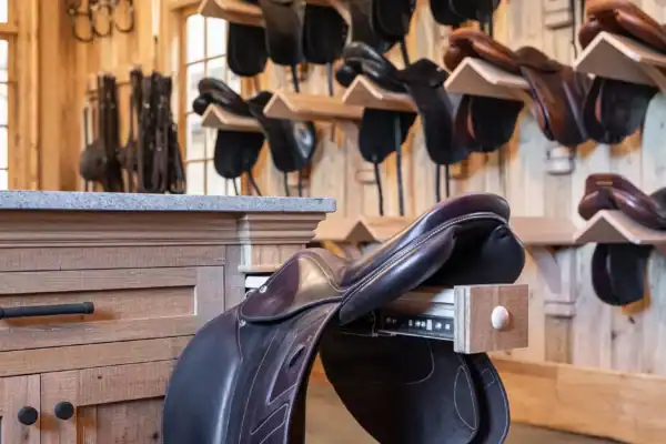 A tack room in a luxury horse barn with wooden walls and multiple horse saddles neatly hung on racks. A brown saddle rests on the edge of a wooden counter in the foreground, all well-lit with natural light.