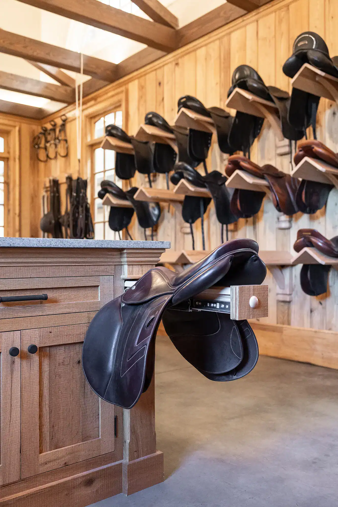 A tack room in a luxury horse barn with wooden walls and multiple horse saddles neatly hung on racks. A brown saddle rests on the edge of a wooden counter in the foreground, all well-lit with natural light.