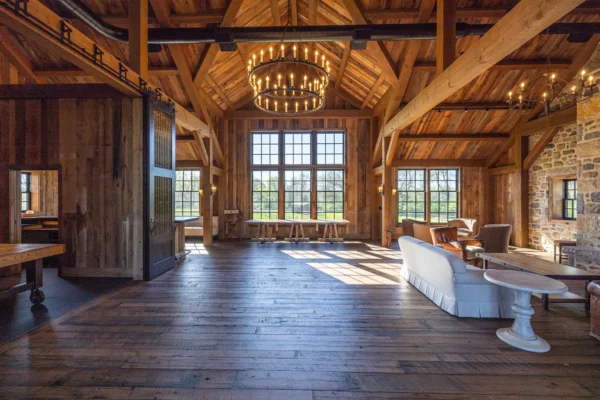Spacious rustic living room in a luxury home with high wooden ceilings, exposed beams, chandeliers, wooden floors, a white sofa, stone wall, and large windows. Natural light streams in for a cozy and inviting atmosphere with countryside views.