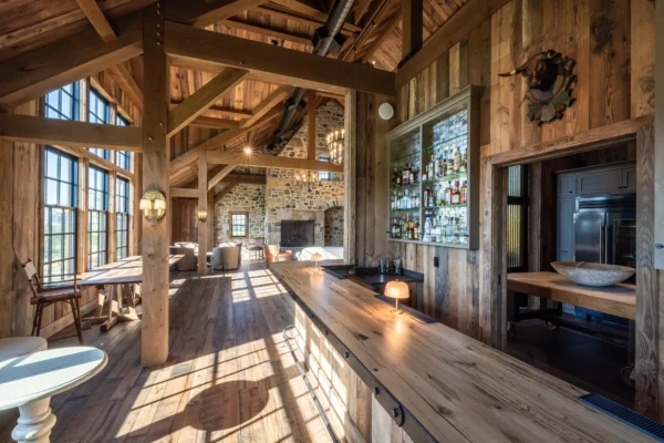 Spacious rustic kitchen and lounge area in a converted bank barn, featuring wooden beams, stone walls, large windows for natural light, a long wooden counter, shelves with bottles, and cozy seating near a fireplace.