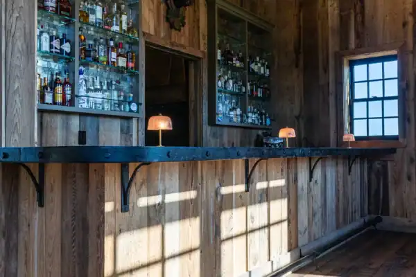 A rustic wooden bar with a metal countertop, shelves stocked with liquor, small lamps on the counter, and sunlight streaming through a window—perfect for gatherings in a luxury horse barn, where charm meets comfort.