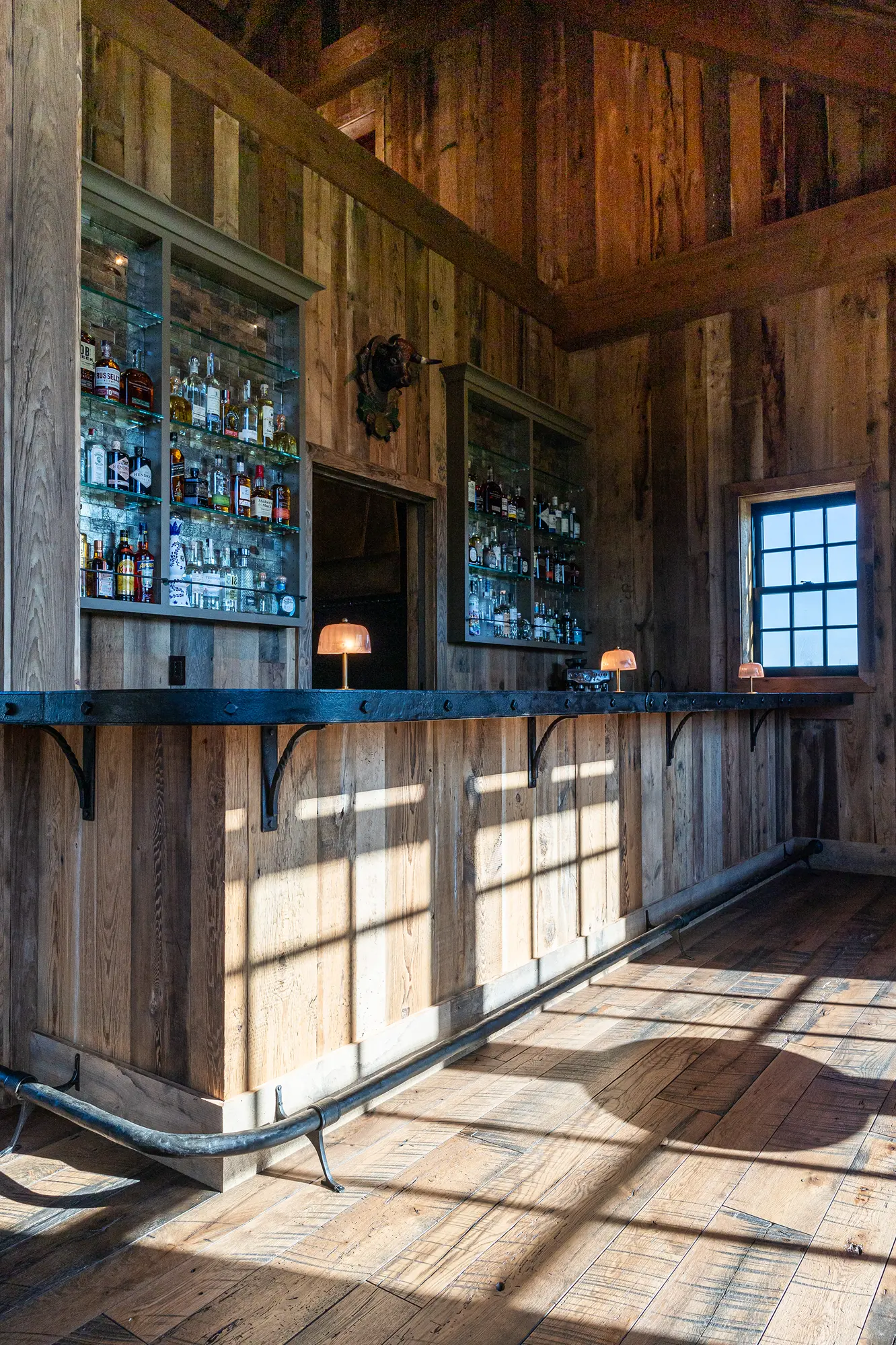 A rustic wooden bar with a metal countertop, shelves stocked with liquor, small lamps on the counter, and sunlight streaming through a window—perfect for gatherings in a luxury horse barn, where charm meets comfort.