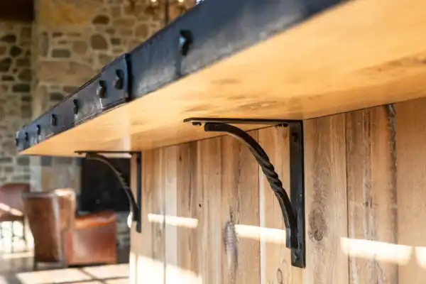 Close-up of a wooden countertop supported by decorative black metal brackets, set against a rustic stone wall reminiscent of a luxury horse barn, with leather chairs blurred in the background.