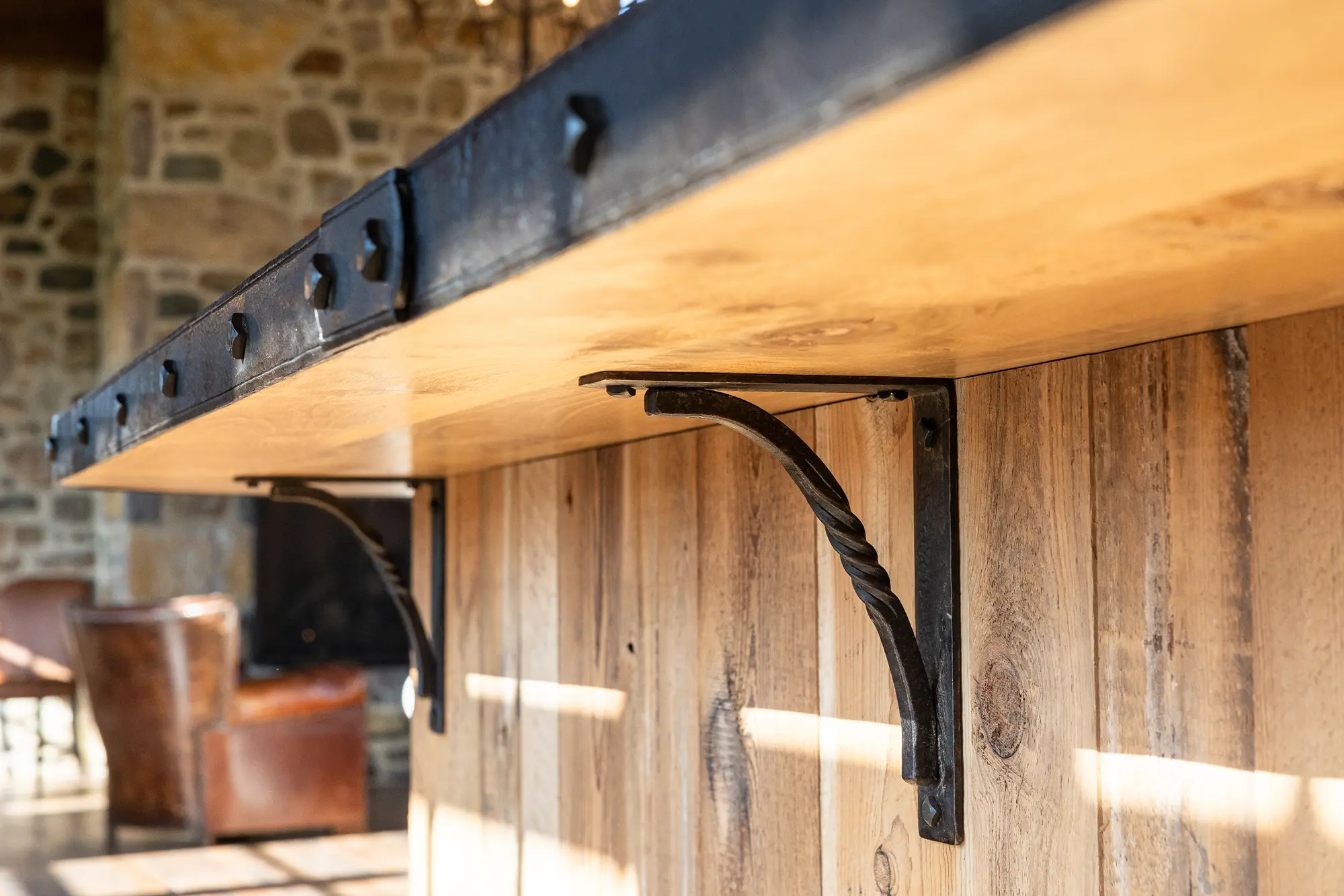 Close-up of a wooden countertop supported by decorative black metal brackets, set against a rustic stone wall reminiscent of a luxury horse barn, with leather chairs blurred in the background.