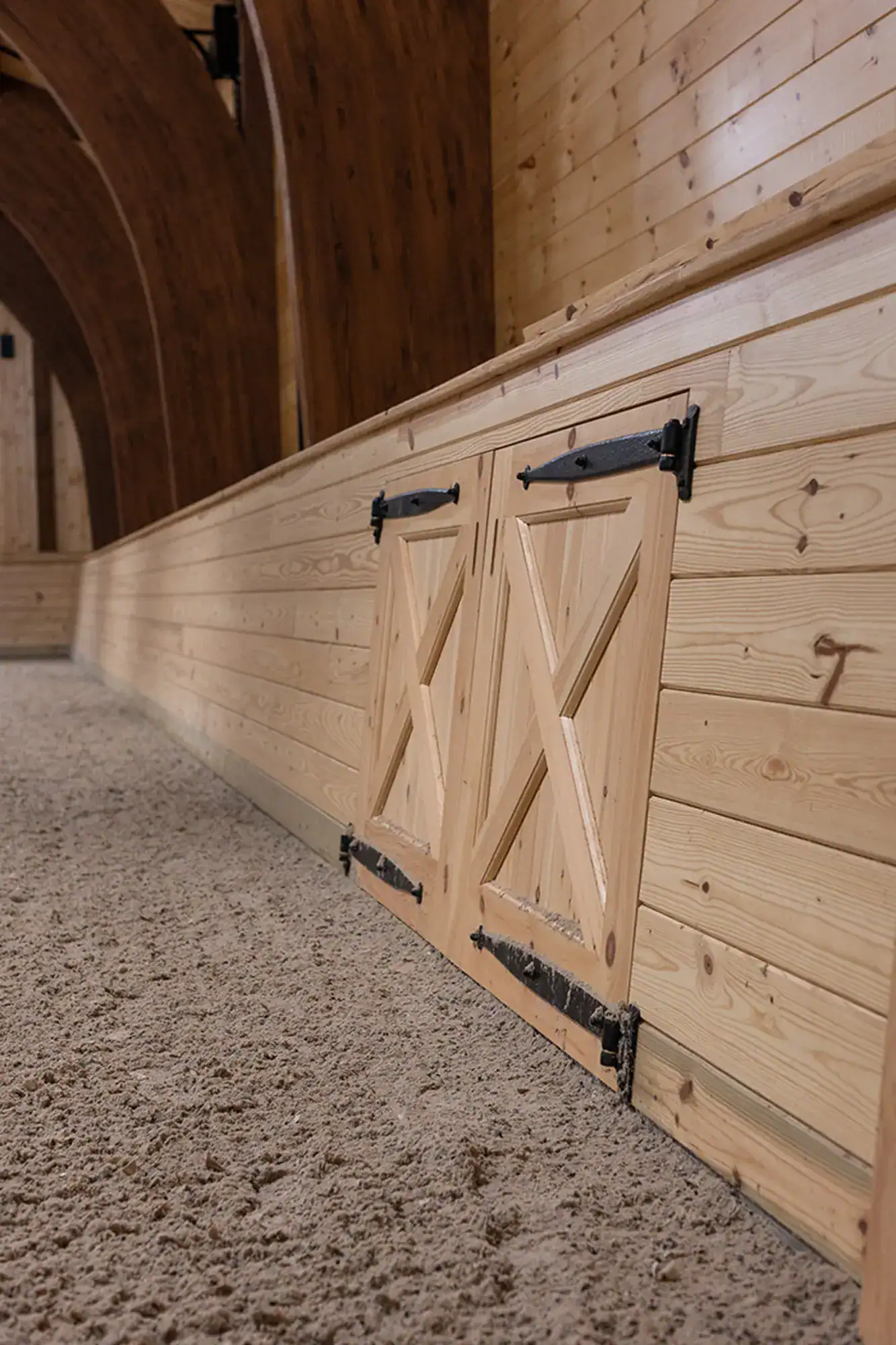 A close-up view of a wooden barn door with black hinges set into a light wood-paneled wall, surrounded by sandy flooring and arched wooden ceiling beams in a luxury horse barn.