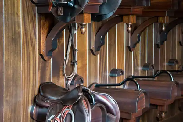 Three brown leather saddles are neatly stored on wooden racks mounted on a wood-paneled wall, adding rustic charm to this luxury horse barn, with stirrups and reins hanging down.