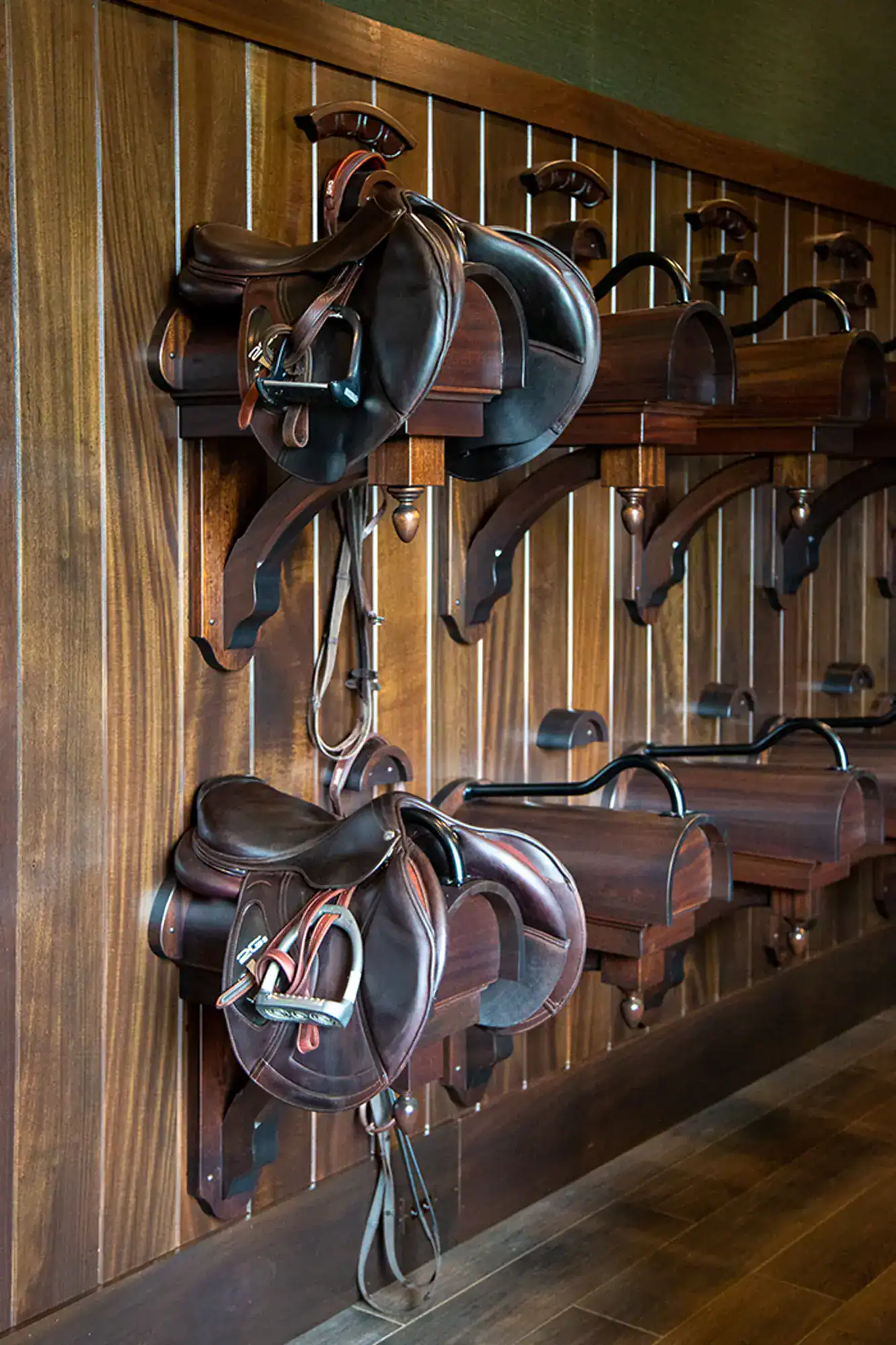 Three brown leather saddles are neatly stored on wooden racks mounted on a wood-paneled wall, adding rustic charm to this luxury horse barn, with stirrups and reins hanging down.