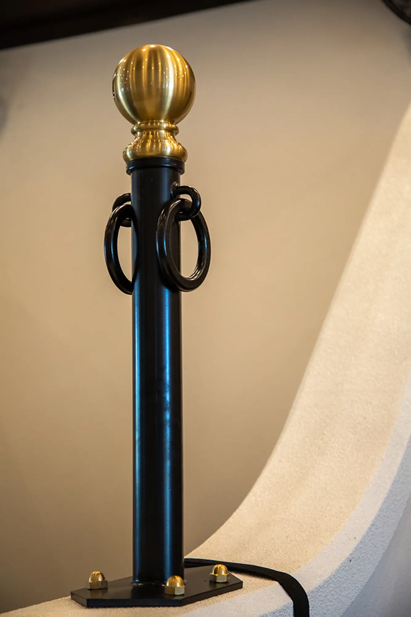 A black metal post with a shiny gold sphere on top and two large circular rings on each side, reminiscent of elegant decor often found in a luxury home, mounted on a light-colored surface against a plain background.