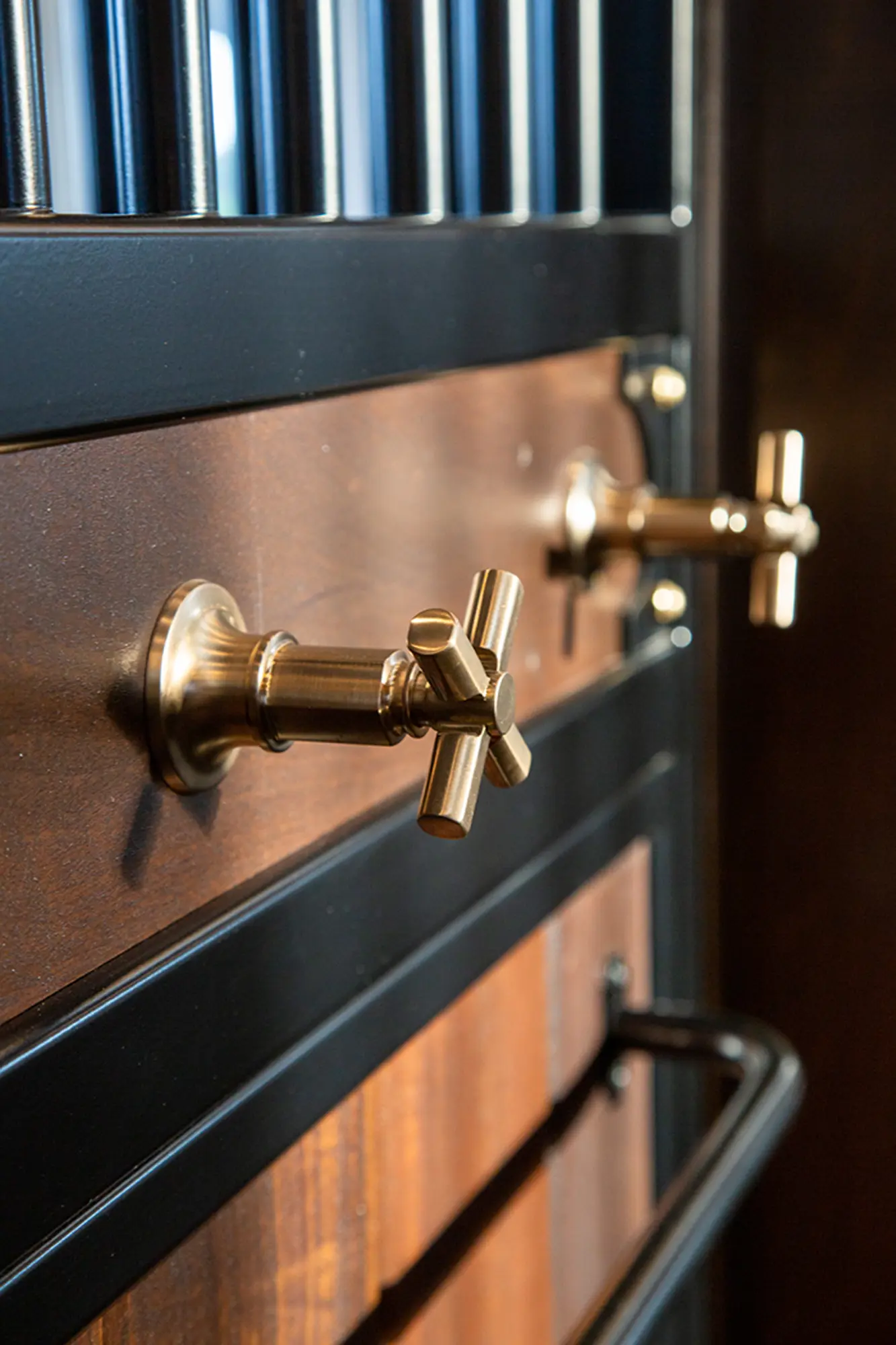 Close-up of two brass water spigots mounted on a dark wood and metal-panel wall in a luxury horse barn, with black metal bars above and a horizontal metal rail below.