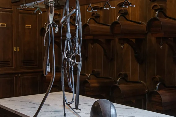 A dark wood tack room in a luxury horse barn features a marble-topped island with drawers, a black riding helmet, and horse tack hanging from a metal rack above. Wooden cubbies and cabinets line the walls in the background.