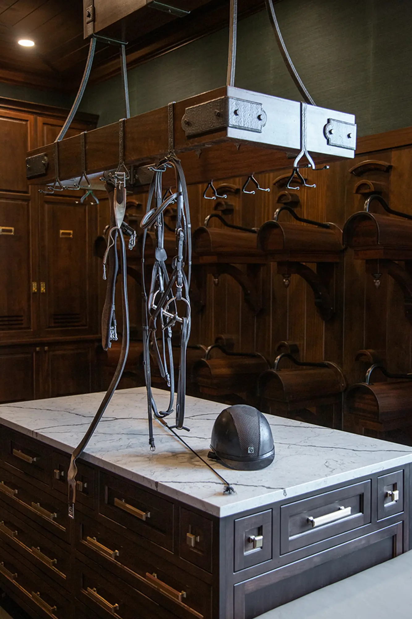 A dark wood tack room in a luxury horse barn features a marble-topped island with drawers, a black riding helmet, and horse tack hanging from a metal rack above. Wooden cubbies and cabinets line the walls in the background.