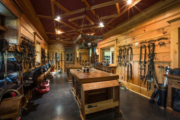 A spacious tack room with wood-paneled walls in a luxury home, a large wooden island in the center, saddles, bridles, and riding gear neatly hanging on racks, and soft lighting from ceiling fixtures.