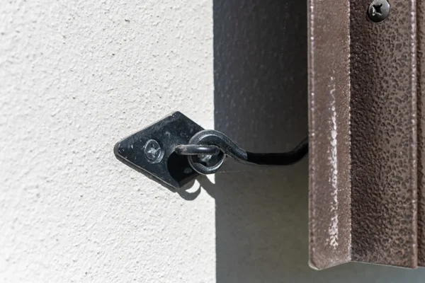 A close-up of a metal hook and eye latch securing a brown shutter to a textured wall in a luxury horse barn. Sunlight casts shadows, highlighting the latch’s detail and the surface textures.