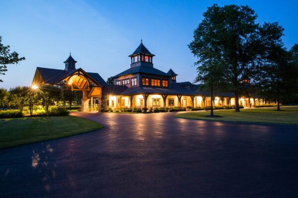 A large, elegant building with tower-like structures and arched windows is warmly lit at dusk, surrounded by trees and a wide driveway under a clear blue sky.