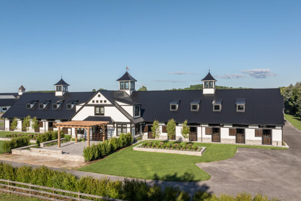 A large, modern barn with a black roof and white walls features three cupolas and a pergola entrance, surrounded by green lawns and trees under a clear blue sky.