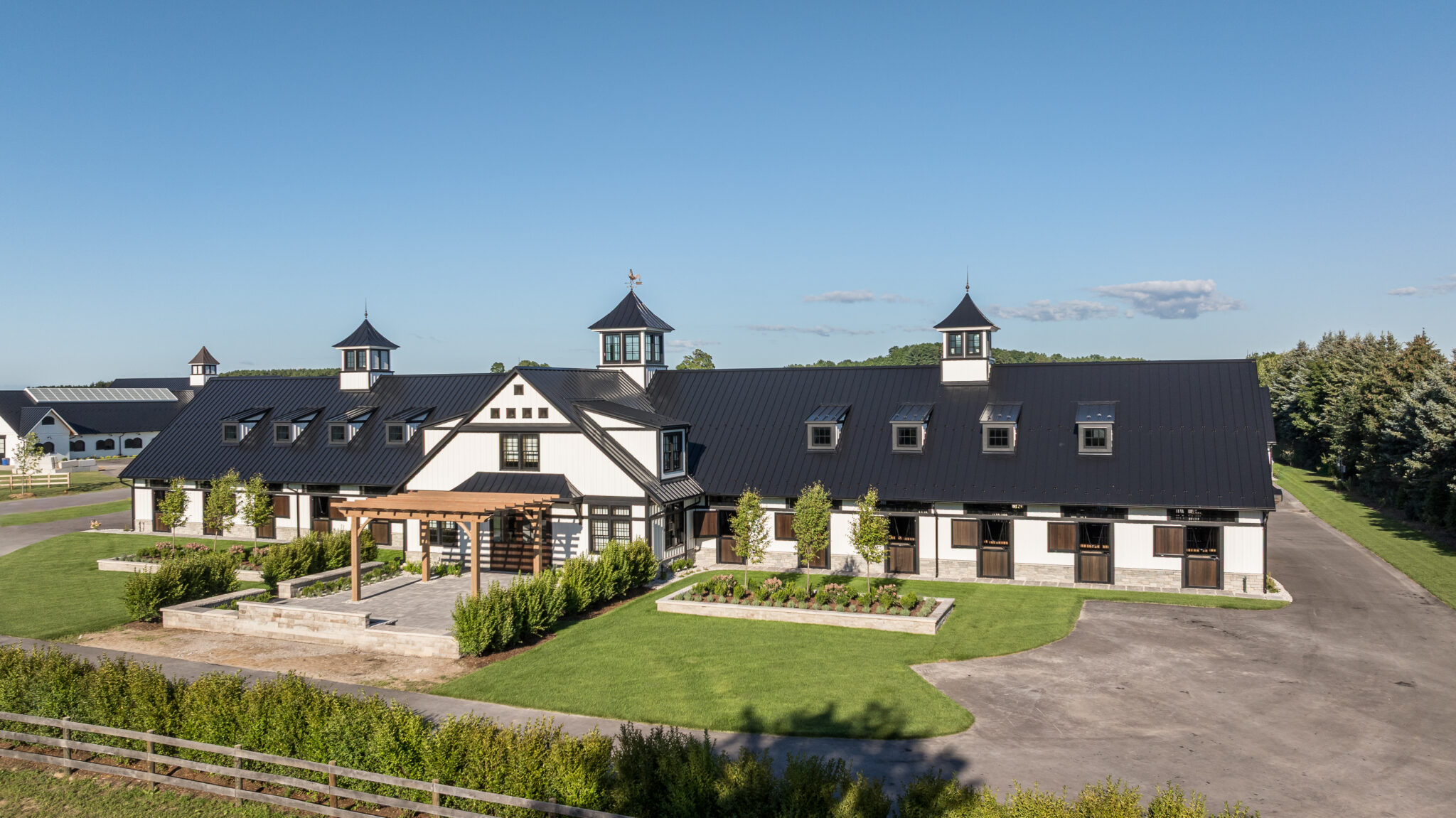 A large, modern barn with a black roof and white walls features three cupolas and a pergola entrance, surrounded by green lawns and trees under a clear blue sky.