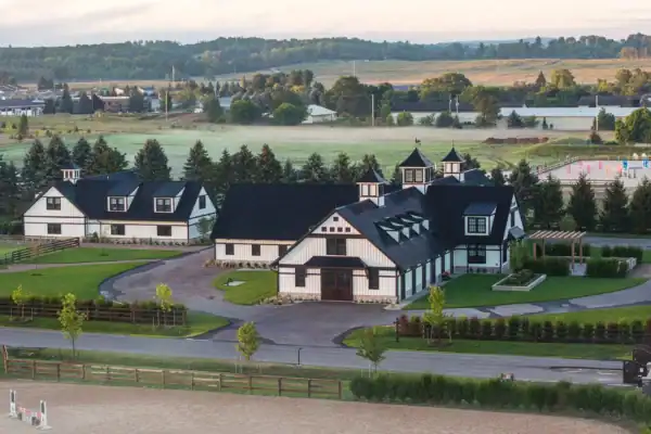 Aerial view of a modern equestrian facility with two large black-and-white barns, fenced pastures, manicured lawns, and a horse riding arena, surrounded by trees and open fields.