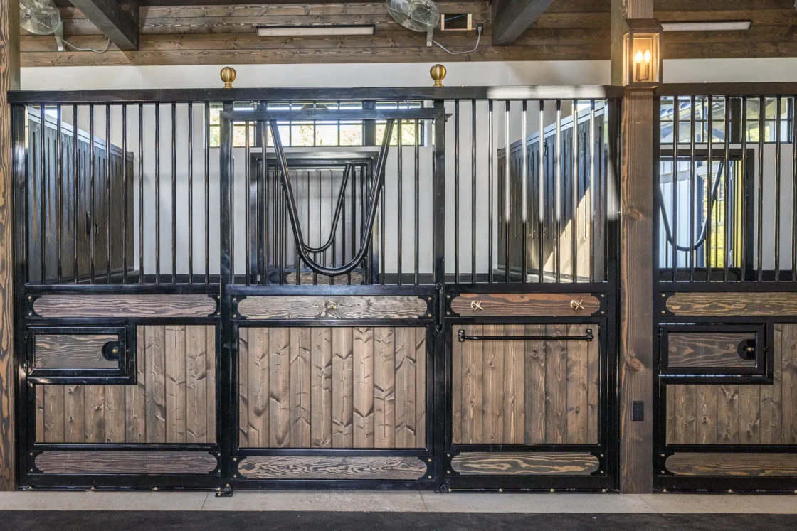 A horse stall with wooden lower panels and black metal bars, featuring sliding doors and decorative accents, inside a well-lit stable with exposed wooden beams.