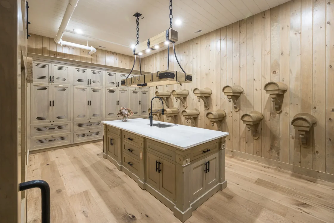 A spacious, rustic mudroom with wood-paneled walls and floors, built-in lockers, an island with a sink, and multiple wooden boot shelves mounted on the right wall. Two industrial light fixtures hang above the island.
