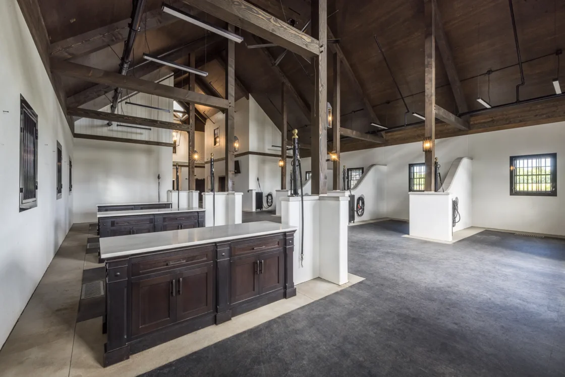 Spacious, modern horse stable with high wooden ceilings, white walls, large windows, and dark wood partitions between stalls. Overhead lights hang from exposed beams, and the floor is black and tan.