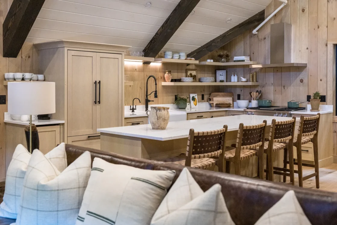 A cozy, modern kitchen with light wood cabinets, open shelves, a white island with four woven chairs, and a farmhouse-style sink. In the foreground, a sofa with plaid pillows faces the kitchen.