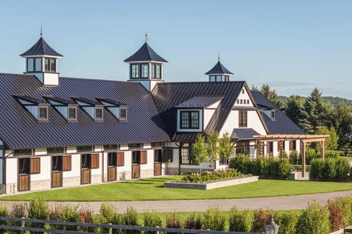 A large, modern barn with white walls, black metal roof, multiple cupolas, and wooden doors, surrounded by manicured lawns, bushes, and trees under a clear blue sky.