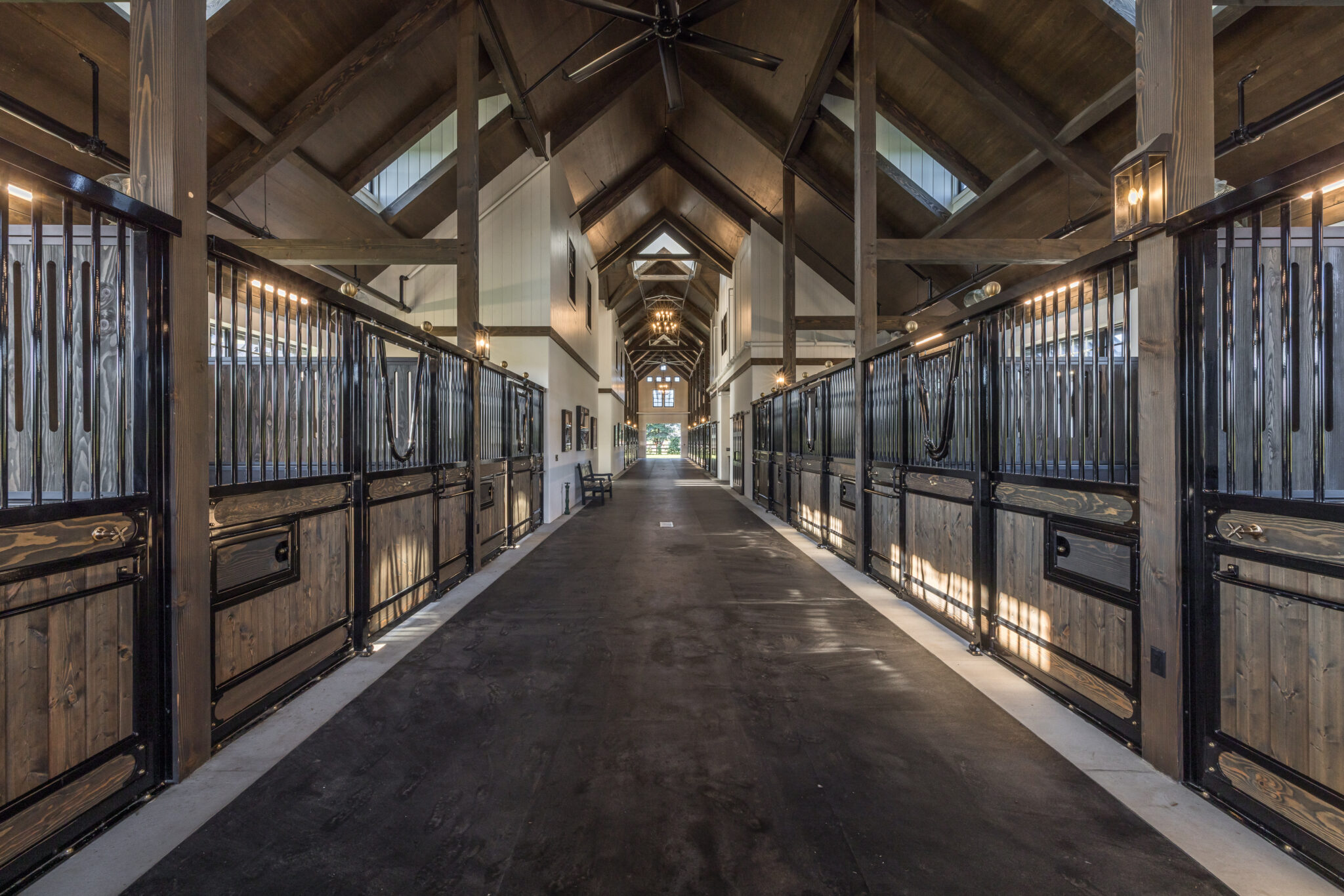 A wide, clean barn aisle with high wooden ceilings, large windows, and rows of empty horse stalls on both sides, illuminated by warm lighting.