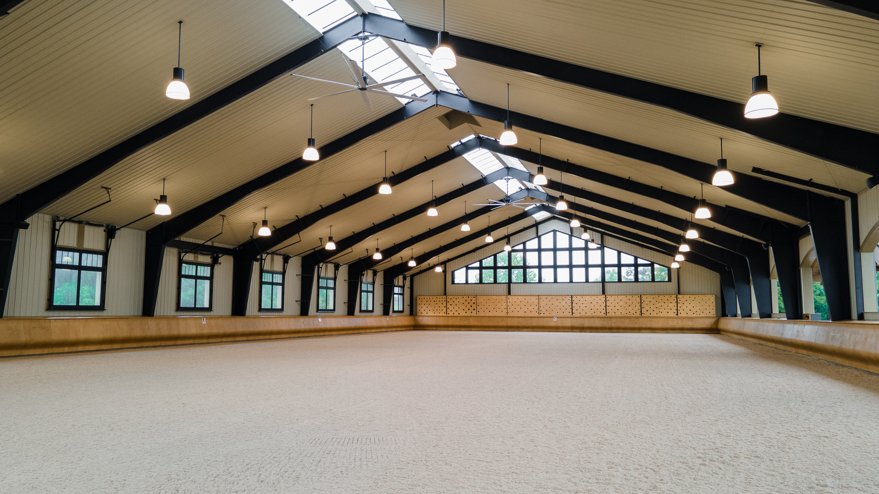 Spacious indoor riding arena with high ceilings, large windows, wooden walls, and bright overhead lights, featuring a clean sand floor and natural light streaming through skylights.